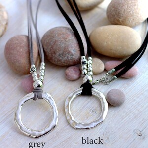May include: Two silver pendant necklaces with a hammered circle design. The necklaces are on a grey and black suede cord with silver beads. The necklaces are displayed on a background of smooth stones.
