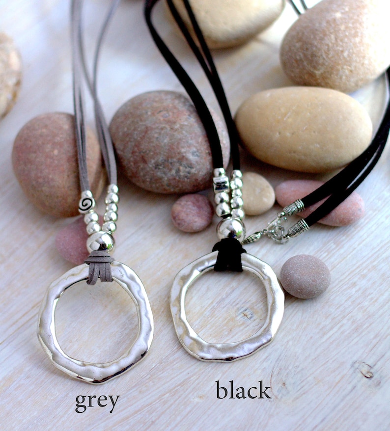 May include: Two silver pendant necklaces with a hammered circle design. The necklaces are on a grey and black suede cord with silver beads. The necklaces are displayed on a background of smooth stones.