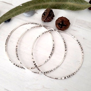 May include: Three silver bangles with etched linear patterns. The thin, circular bracelets are displayed on a white surface, with natural elements in the background. The bangles are a simple, elegant jewelry item.