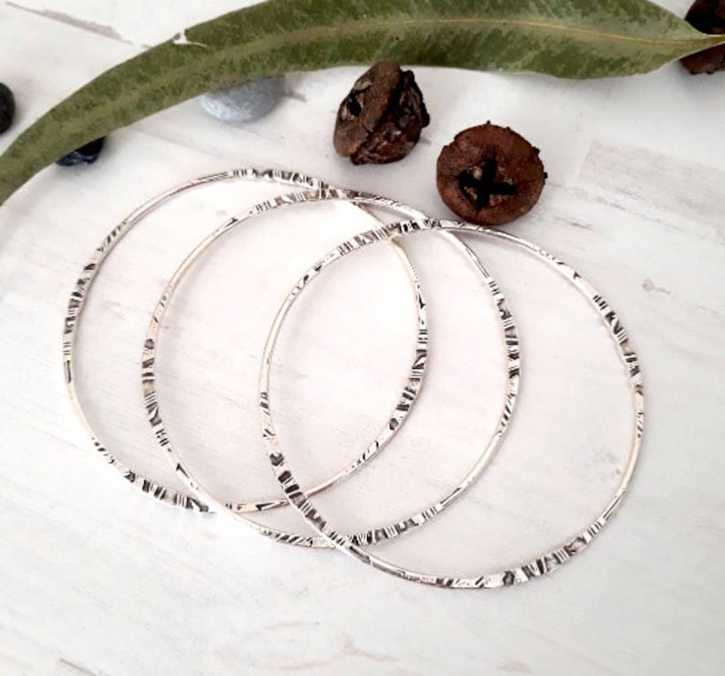 May include: Three silver bangles with etched linear patterns. The thin, circular bracelets are displayed on a white surface, with natural elements in the background. The bangles are a simple, elegant jewelry item.