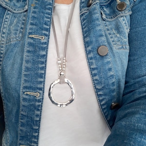 May include: A silver necklace with a large, hammered circle pendant and gray suede cord. The necklace is worn over a white t-shirt and a denim jacket.