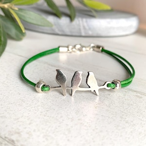 Op de afbeelding: Een armband met een felgroen leren koord en een zilverkleurige metalen bedel die drie kleine vogels op een tak voorstelt. Het ontwerp bevat twee kleine zilverkleurige kralen aan weerszijden van de vogelbedel en een zilverkleurige karabijnsluiting.