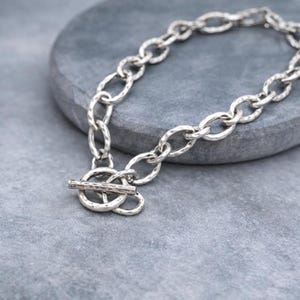 May include: A silver-toned chain bracelet featuring oval-shaped links with a textured, hammered finish. The bracelet is secured with a toggle clasp consisting of a circular ring and a straight bar, both matching the hammered metal design.