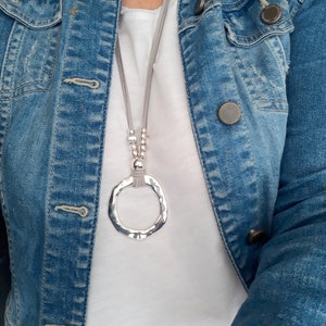 May include: A silver pendant necklace with a large, hammered circle pendant hangs from a gray suede cord. The necklace is worn over a white t-shirt and a denim jacket.