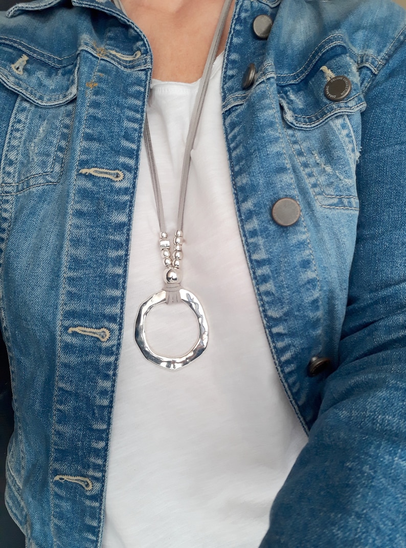 May include: A silver pendant necklace with a large, hammered circle pendant hangs from a gray suede cord. The necklace is worn over a white t-shirt and a denim jacket.