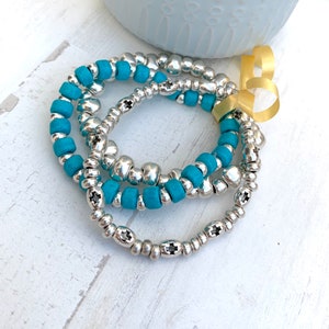 Op de afbeelding: Drie turquoise en zilveren kralen armbanden gestapeld op een wit oppervlak. De armbanden zijn gemaakt van kleine, ronde kralen met een kruisontwerp op de zilveren kralen.
