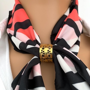 Könnte beinhalten: Goldener Schalring mit einem dekorativen, durchbrochenen Design. Der Ring hält einen schwarz-weiß gestreiften Schal mit rosa und roten Akzenten.