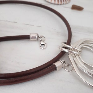 Peut inclure: Un collier en cordon de cuir marron avec un pendentif argenté composé de plusieurs cercles entrelacés. Le collier a un fermoir argenté et deux fermoirs à barillet argentés.
