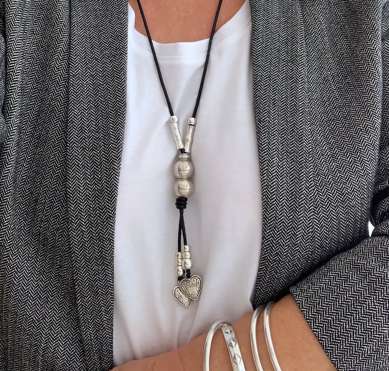 May include: A long black leather necklace with silver beads and heart charms. The necklace is worn by a person wearing a white shirt and a gray blazer.