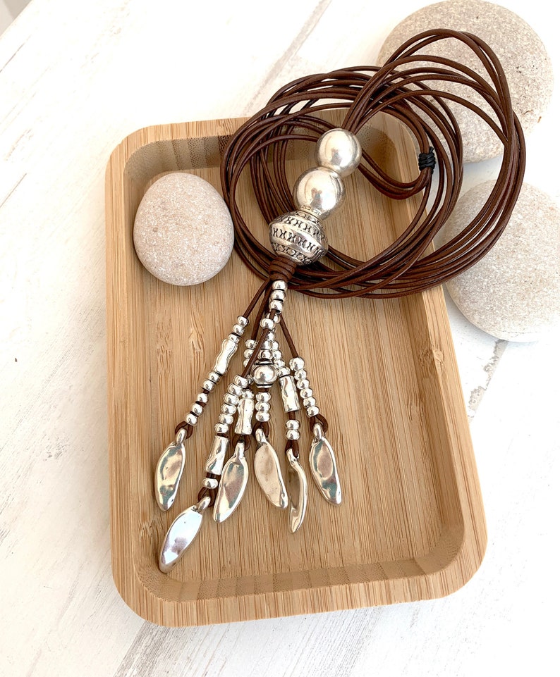 K&ouml;nnte beinhalten: Eine lange braune Lederkette mit silberfarbenen Perlen und blattf&ouml;rmigen Anh&auml;ngern. Die Kette wird auf einem Holztablett mit zwei glatten Steinen pr&auml;sentiert. Die Kette hat einen Bohemian-Stil.