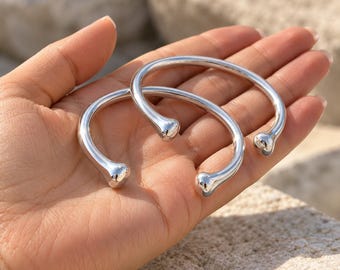 May include: Two polished silver-tone cuff bracelets with a smooth, rounded finish. Each bracelet features an open design with a small, rounded ball terminal at both ends, creating a clean, reflective surface that catches the light.