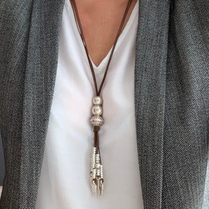 K&ouml;nnte beinhalten: Eine lange Halskette mit braunem Lederband und silberfarbenen Perlen und einer Quaste. Die Halskette wird &uuml;ber einem wei&szlig;en Oberteil und einem grauen Blazer mit Fischgr&auml;tenmuster getragen.