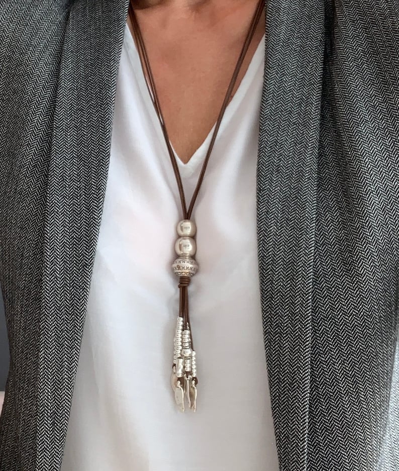 K&ouml;nnte beinhalten: Eine lange Halskette mit braunem Lederband und silberfarbenen Perlen und einer Quaste. Die Halskette wird &uuml;ber einem wei&szlig;en Oberteil und einem grauen Blazer mit Fischgr&auml;tenmuster getragen.