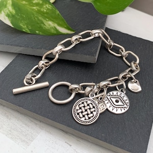 May include: Silver charm bracelet with a toggle clasp and multiple charms, including a woven circle, an evil eye, and a heart.