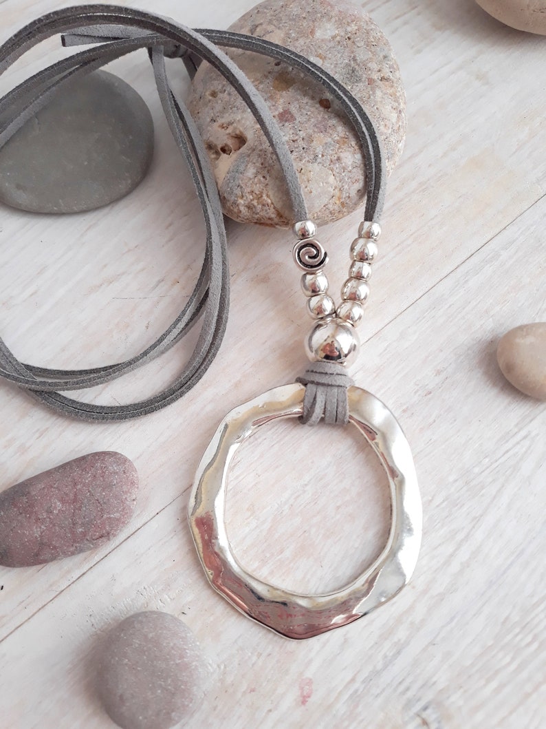 May include: A silver pendant necklace with a large, hammered, circular pendant. The necklace is made with a gray suede cord and silver beads.
