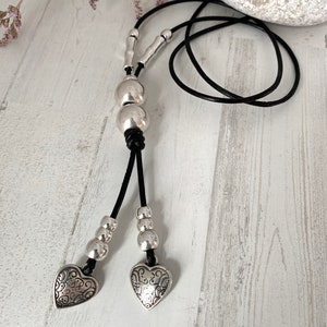 May include: A black leather cord necklace with silver beads and two silver heart charms. The hearts have a decorative design.
