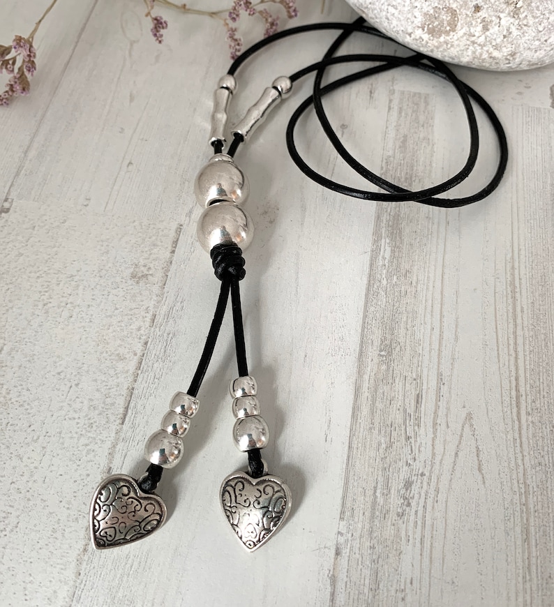 May include: A black leather cord necklace with silver beads and two silver heart charms. The hearts have a decorative design.