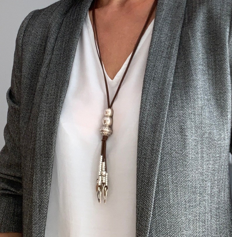 K&ouml;nnte beinhalten: Eine lange braune Lederkette mit silberfarbenen Metallperlen und einem dekorativen Anh&auml;nger. Die Kette wird &uuml;ber einem wei&szlig;en Oberteil und einem grauen Blazer getragen.