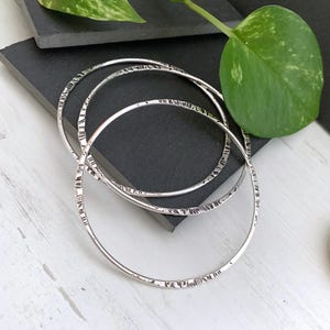 May include: Three silver bangles with textured detailing. The bangles are circular and have a hammered or etched pattern around the bands. The jewelry is displayed on a dark gray surface with a green leaf in the background.