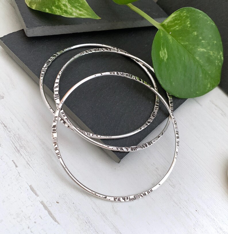 May include: Three silver bangles with textured detailing. The bangles are circular and have a hammered or etched pattern around the bands. The jewelry is displayed on a dark gray surface with a green leaf in the background.
