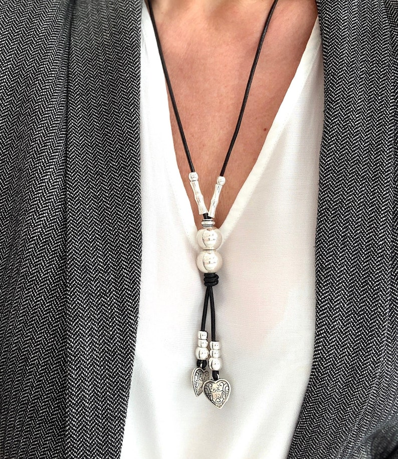 May include: A black leather necklace with silver beads and two heart charms. The necklace is worn by a person wearing a white shirt and a gray blazer.