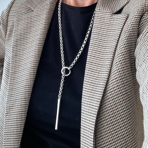 Könnte beinhalten: Eine silberne Kettenkette mit einem langen, dünnen Anhänger und einem runden Ring oben. Die Kette wird über einem schwarzen Shirt und einem braunen und weißen gemusterten Blazer getragen.