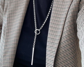 May include: A silver-toned necklace with a long, vertical bar pendant. The necklace features a chain with a circular ring and a bar pendant. The person is wearing a black shirt and a brown and white plaid blazer.