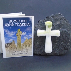 Puede incluir: Una cruz de mármol blanco con vetas verdes, tallada en mármol de Iona, un tipo de serpentina que se encuentra en la isla de Iona, Escocia. La cruz se exhibe sobre una roca negra con un pequeño folleto que dice "Scottish Iona Marble" y "A wee piece of the sacred isle".
