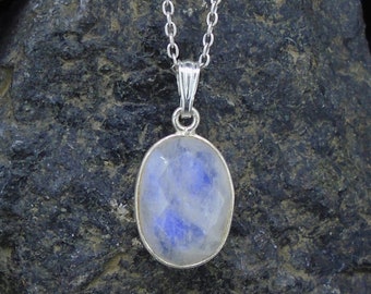 May include: A silver necklace featuring an oval-shaped moonstone pendant with a faceted surface. The pendant is suspended from a delicate silver chain, showcasing a translucent stone with subtle blue iridescent flashes.