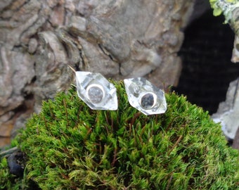 May include: A pair of stud earrings featuring clear, multifaceted, geometric crystal points. Each earring has a small, circular, dark gray metallic center set within the transparent, angular crystal structure.