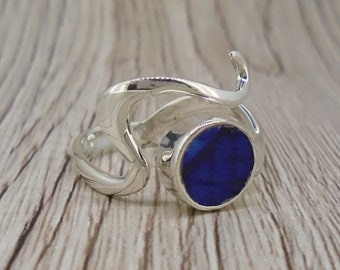 May include: A silver ring with a blue gemstone set in a circular bezel. The ring band is a twisted, abstract design.