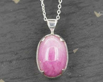 May include: A silver pendant necklace featuring a smooth, oval-shaped pink gemstone set in a polished silver prong setting. The pendant hangs from a delicate silver chain with small, uniform circular links, creating a simple and elegant piece.