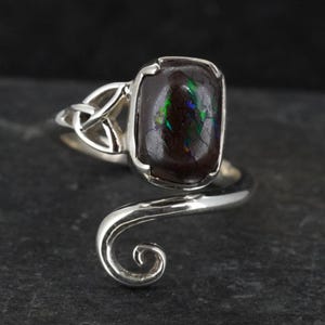 Op de afbeelding: Zilveren ring met een donkere, rechthoekige edelsteen met iriserende groene en blauwe flitsen. De ring heeft een Keltisch knoopontwerp en een sierlijke band. De steen is ongeveer 1,5 cm lang.
