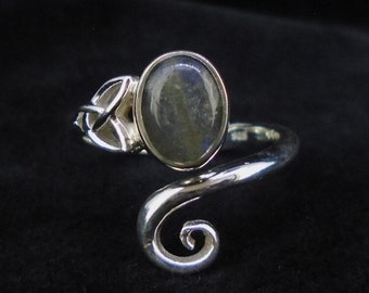 Può includere: Un anello d'argento con una fascia a spirale e una pietra rotonda blu-grigia iridescente incastonata in un design a nodo celtico.