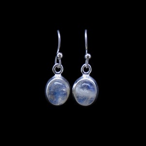 May include: A pair of silver drop earrings featuring oval-shaped moonstones with a translucent, iridescent blue sheen. Each stone is set in a smooth silver bezel, suspended from a simple silver hook wire with a small decorative silver bead.