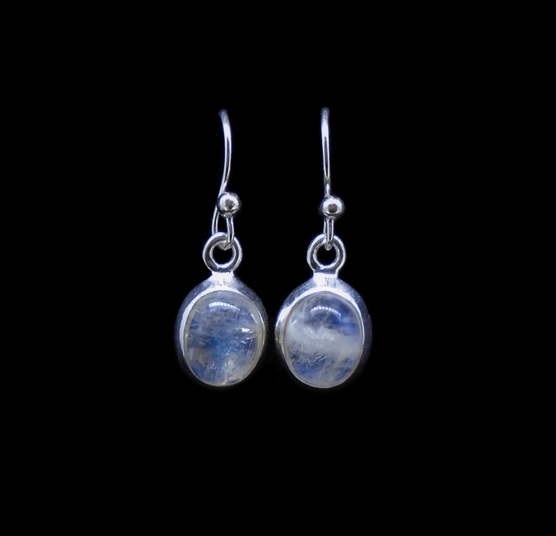 May include: A pair of silver drop earrings featuring oval-shaped moonstones with a translucent, iridescent blue sheen. Each stone is set in a smooth silver bezel, suspended from a simple silver hook wire with a small decorative silver bead.