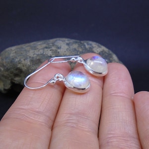 May include: A pair of silver drop earrings featuring oval-shaped moonstones with a translucent, iridescent sheen. Each stone is set in a smooth silver bezel, attached to a simple silver hook wire with a small decorative silver bead.