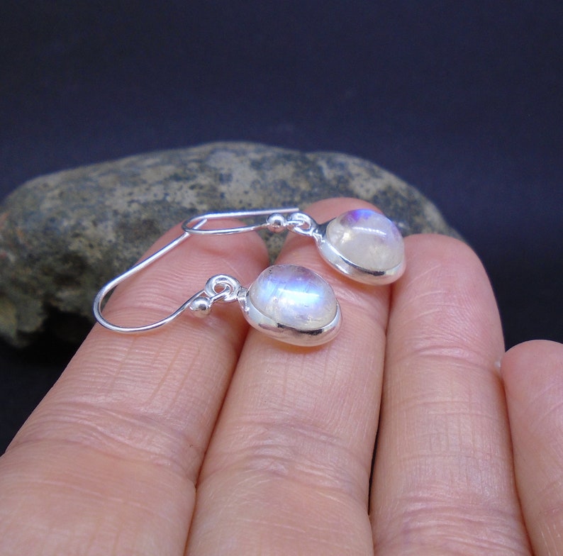May include: A pair of silver drop earrings featuring oval-shaped moonstones with a translucent, iridescent sheen. Each stone is set in a smooth silver bezel, attached to a simple silver hook wire with a small decorative silver bead.