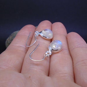 May include: A pair of silver drop earrings featuring round, translucent moonstones with a subtle blue sheen. Each earring is set in a smooth silver bezel with a small silver bead accent at the top, connected to a simple silver hook wire.