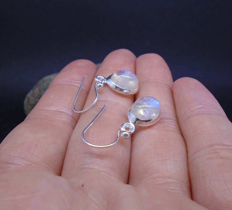 May include: A pair of silver drop earrings featuring round, translucent moonstones with a subtle blue sheen. Each earring is set in a smooth silver bezel with a small silver bead accent at the top, connected to a simple silver hook wire.