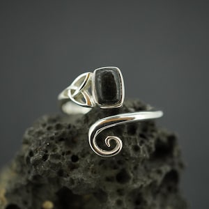 May include: A silver bypass ring featuring a rectangular black stone set in a polished metal frame. One side of the band displays a triquetra knot design, while the other side terminates in a smooth, tapered spiral curl that curves outward.