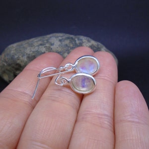 May include: A pair of silver drop earrings featuring oval-shaped moonstones with a translucent, iridescent sheen. The earrings are designed with simple silver hooks for attachment, showcasing a polished metal setting that frames each smooth, rounded stone.
