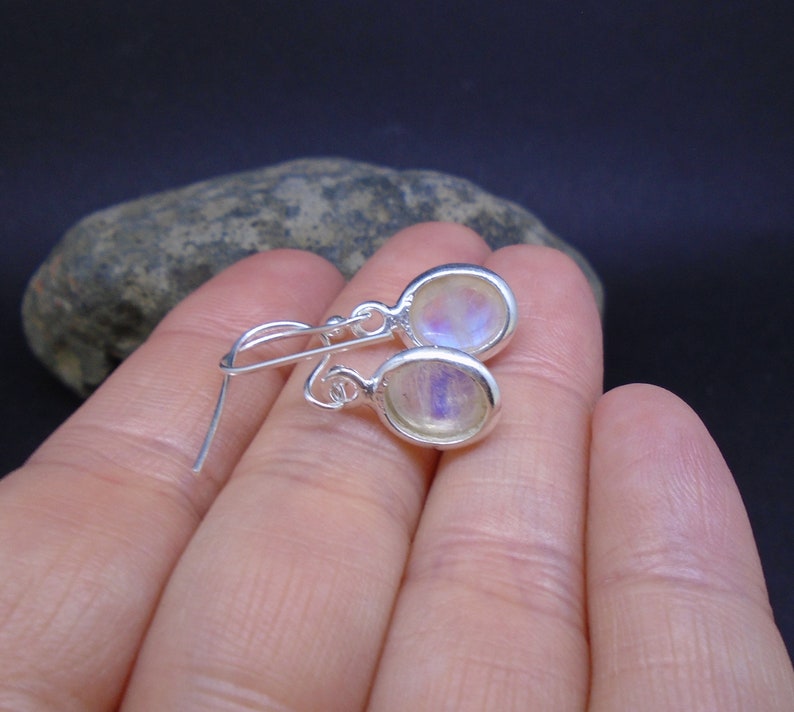 May include: A pair of silver drop earrings featuring oval-shaped moonstones with a translucent, iridescent sheen. The earrings are designed with simple silver hooks for attachment, showcasing a polished metal setting that frames each smooth, rounded stone.