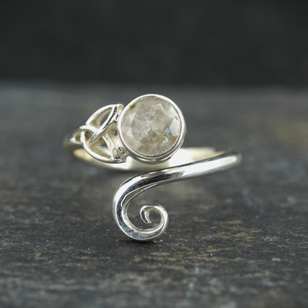 May include: A silver ring with a round, faceted, light-colored gemstone. The ring features a Celtic knot design on one side and a swirling, open design on the other. The ring is set against a dark background.