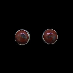 May include: A pair of round stud earrings featuring polished, deep red, mottled stones set in simple silver-toned metal bezels. The stones display natural variations in color with dark brown and black inclusions throughout the smooth, domed surfaces.
