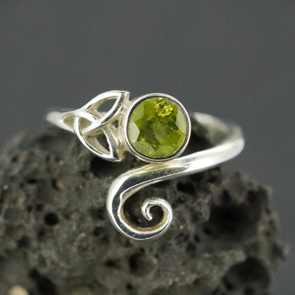 May include: A silver ring featuring a round, faceted lime green gemstone set in a bezel. One side of the band displays an openwork triquetra knot design, while the other side terminates in a smooth, polished silver spiral that curves outward.