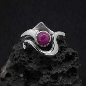 Può includere: Un anello in argento con una vibrante gemma di rubino rotonda. L'anello presenta un design unico e fluido con elementi curvi e angolari. Il rubino è incastonato in una lunetta e l'anello è esposto su una superficie scura e strutturata.