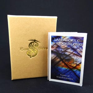 May include: A tan rectangular box with a gold embossed Two Skies logo next to a small white booklet featuring a blue and purple labradorite and moonstone graphic with the text Labradorite & Moonstone Gems of transformation.