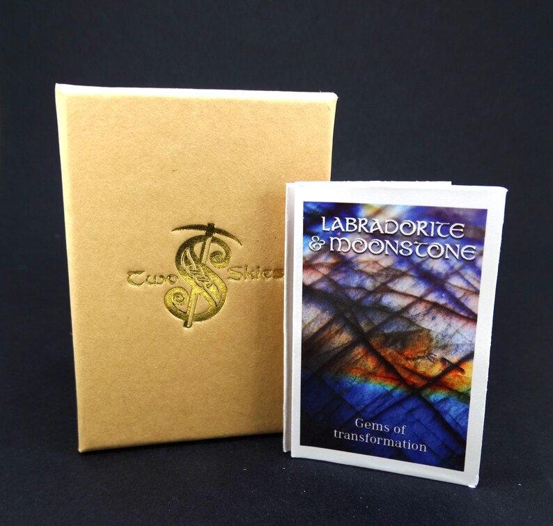 May include: A tan rectangular box with a gold embossed Two Skies logo next to a small white booklet featuring a blue and purple labradorite and moonstone graphic with the text Labradorite & Moonstone Gems of transformation.