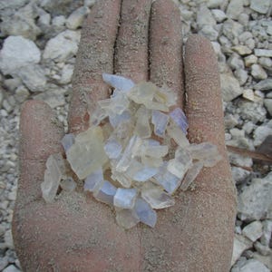 May include: A collection of small, translucent, and semi-opaque raw mineral fragments in shades of pale blue, white, and clear. The stones have irregular, jagged edges and a smooth, glassy texture, appearing as unpolished, natural crystal pieces.